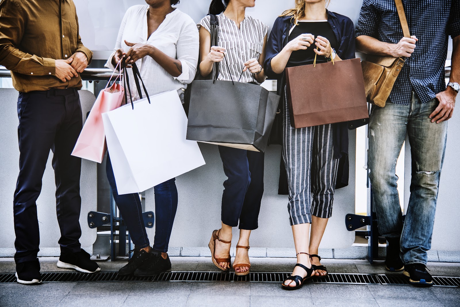 Five people with their heads cut out of the picture stand against a wall while holding shopping bags